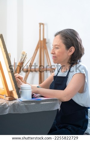 Happy female artist near workplace, mature female artist in black apron standing with crossed arms near table with brushes and assorted paints while looking at camera in studio.