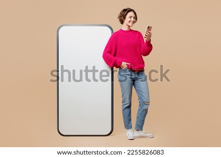 Full body smiling fun young woman wear pink sweater big huge blank screen mobile cell phone with workspace copy space mockup area using hold smartphone isolated on plain pastel light beige background