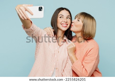 Elder fun parent mom with young adult daughter two women together wear casual clothes doing selfie shot on mobile cell phone hug cuddle kiss isolated on plain blue cyan background. Family day concept