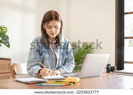 Female fashion artist designing new fashion and sketching or drawing model with dress in notebook while working with laptop in workshop studio.