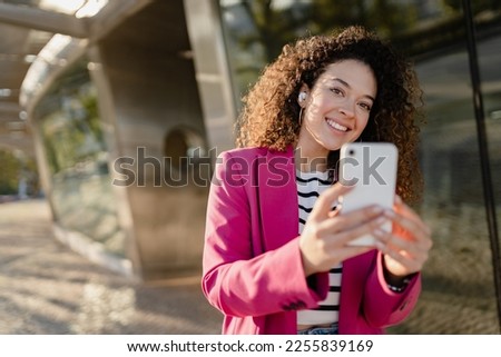 pretty curly woman in city street in stylish pink jacket, student education, using smartphone, taking selfie picture, making photo