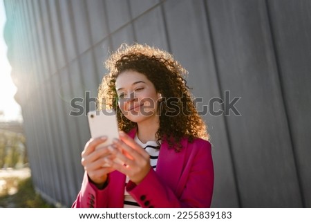 pretty curly smiling woman walking in city street in stylish pink jacket, using smartphone, taking selfie picture, making photo