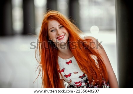a red-haired girl in a light skirt; a very cheerful girl with long red hair; lips painted with red lipstick; sits on a bench and has a good mood; poses for a photographer; street photo session  Royalty-Free Stock Photo #2255848207