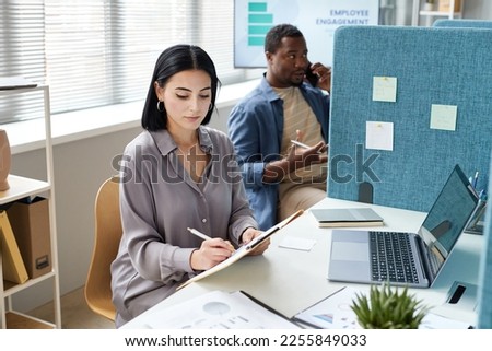 Portrait of young businesswoman writing on clipboard while working in office cubicle