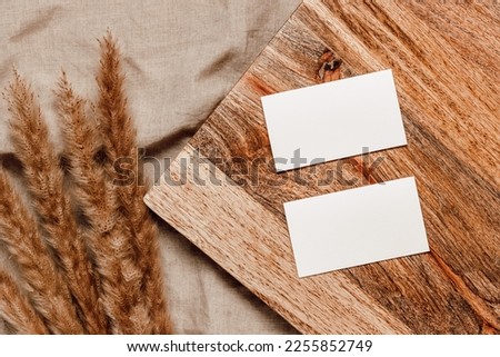 Two blank paper mockup cards on wooden stand with reed or pampas grass on linen beige cloth background. Minimal paper business card, social media, invitation and blog template, aesthetic style. 