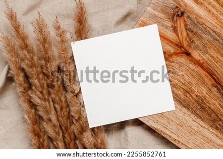 Blank paper mockup card on wooden stand with reed or pampas grass on linen beige cloth background. Minimal paper sheet card for invitation, social media, business and blog template, aesthetic style. 