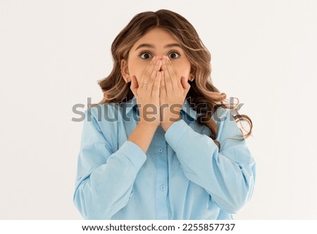 Young shocked woman closed her mouth with crossed hands and wide opened eyes. Horizontal photo of european female isolated on white background with expression of secrecy and fear on her face.