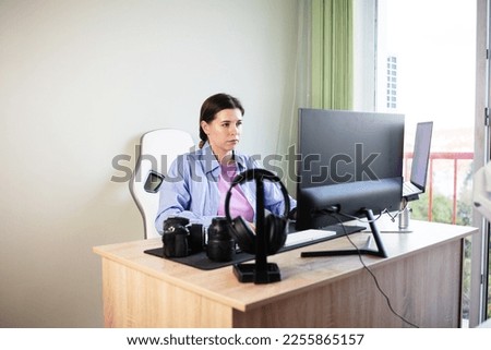 Photographer in casual clothing, sitting at her desk, uses a desktop computer in retouching. Desktop with two monitors, keyboard, camera, diary, palette for designer, retoucher, photographer