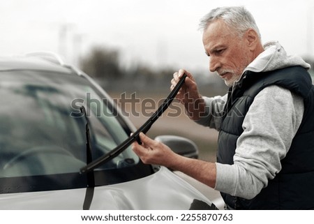 Caucasian senior 60-70 years old man driver holding new brand windscreen wipers and replacing them, mounting on the windshield car on outdoors parking lot of a car station. Car service concept  Royalty-Free Stock Photo #2255870763
