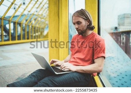 Surprised thoughtful freelancer in casual wear working on startup using laptop looking for decision of problem in covered pedestrian bridge Royalty-Free Stock Photo #2255873435