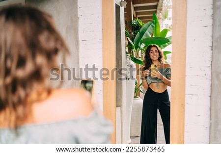Optimistic stylish ethnic female in trendy wear smiling while taking photo on mobile phone and looking at mirror with joy