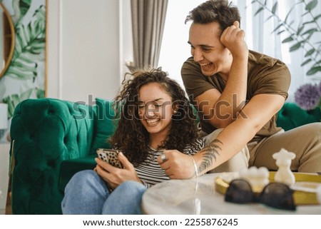 caucasian woman using mobile phone for online sms chat or internet browsing while her boyfriend or husband adult male sit beside at home in bright room real people couple leisure concept