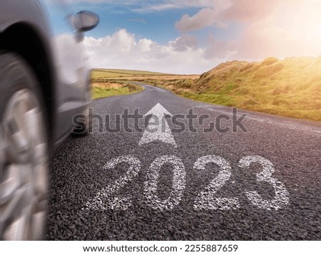 Sign 2023 on small asphalt country road and and arrow and side of a car in motion blur. Travel and explore concept. Warm sunny day, sun flare. Green hill on a side. Blue cloudy sky.