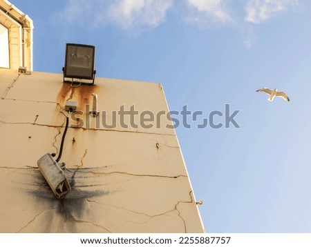 Damaged CCTV camera hanging on a cable on yellow wall, old flood light above the camera. Front element is broken and there is sign of explosion. Seagull in blue cloudy sky. War or unsafe area.
