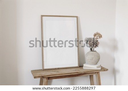 Neutral artistic still life. Blank picture frame mockup on old wooden bench in sunlight. Dry hydrangea flowers in textured vase. White wall background. Empty copyspace. Elegant boho interior.  