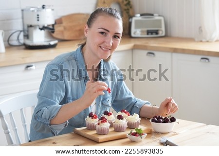 A young woman decorates muffins with white cream and berries in her kitchen. Food and people concept.