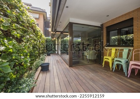 Terrace of a house on the ground floor with hedges and decorative plants, acacia floors, glass partitions and colored metal chairs