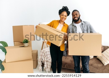 Moving to a new apartment. Happy newlyweds bought their first home, stand in the living room among the boxes, hold cardboard boxes in their hands, prepare to unpack things, look at the camera, smile