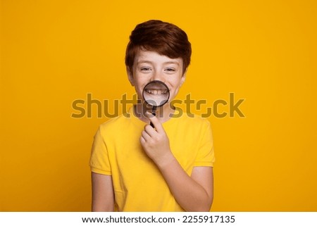Closeup studio photo portrait of satisfied caucasian boy holding transparent magnifying glass on his mouth zooms in his smile and teeth isolated over yellow