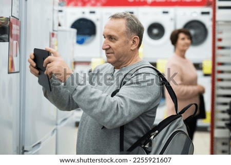 Mature man standing in salesroom of appliance store and photographing refrigerator with his smartphone.