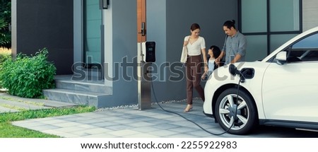 Progressive young parents and daughter with electric vehicle and home charging station. Green and clean energy from electric vehicles for healthy environment. Eco power from renewable source at home. Royalty-Free Stock Photo #2255922983