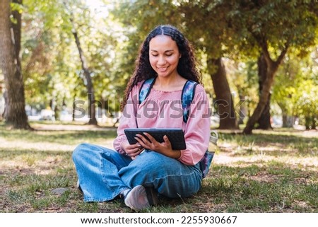 Latin university student woman smiling and using a tablet sitting outside in a park on the grass