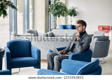 Confident young man in formal suit and glasses, sitting in an armchair in the VIP lounge of a business office, checking his CV resume while waiting for a job interview. People and recruitment concept Royalty-Free Stock Photo #2255963855