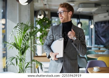 Stylish young businessman in formal suit and glasses smiling looking away, holding a laptop in hands while standing in the modern office interior. Successful purposeful people and business concept Royalty-Free Stock Photo #2255963919