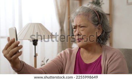 Asian old lady with presbyopia is holding her mobile phone far from eyes as she is having trouble reading prints on the screen clearly in the living room at home Royalty-Free Stock Photo #2255998649
