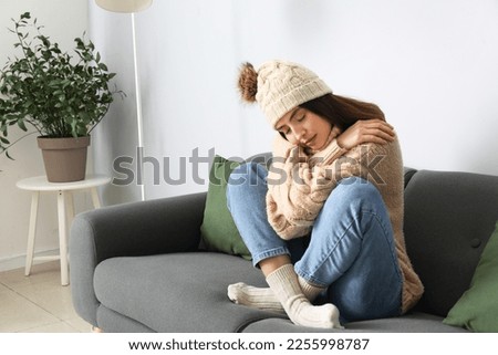 Frozen young woman in winter clothes sitting on sofa at home