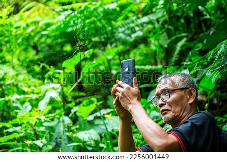 an old man is learning to take pictures using his cell phone to take pictures insects or spiders hanging in front of him in a park where recreation