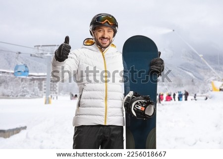 Young man with snowboard gesturing thumbs up and smiling in a ski resort