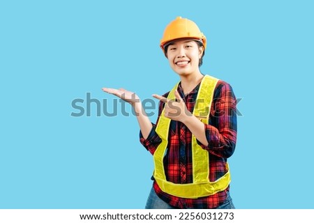 Portrait Young female engineer in plaid shirt wear safety jacket and helmet standing open palm and point finger gesture with smile, isolated on blue background, copy space to insert your advertising