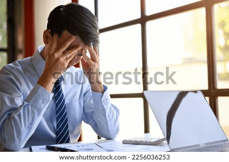 Stressed millennial Asian businessman sits at his desk, worried about his business's financial investment outcome, getting trouble from clients, or dissatisfied with work's performance. Royalty-Free Stock Photo #2256036253