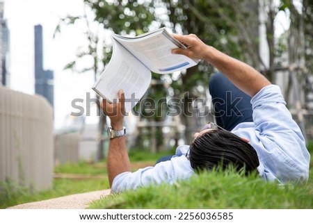 Side view of handsome man reading book in grass under tree with glasses. Relax in summer time holiday laying on the grass field comfortable feeling. Soft focus