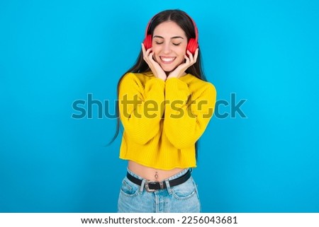 Pleased young brunette girl wearing yellow knitted sweater against blue wall enjoys listening pleasant melody keeps hands on stereo headphones closes eyes. Spending free time with music Royalty-Free Stock Photo #2256043681