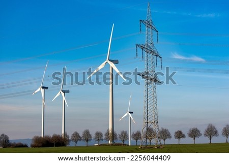 The wind turbines of a wind farm for the production of sustainable energy on the horizon against an evening blue, slightly cloudy sky with a power pole Royalty-Free Stock Photo #2256044409