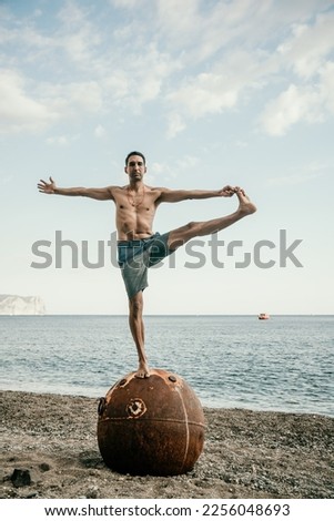 Man doing yoga exercise outdoors standing on old rusty floating marine mine on the beach with rocky shore and sea background. Healthy lifestyle, pollution, nature protection, war and peace concept.