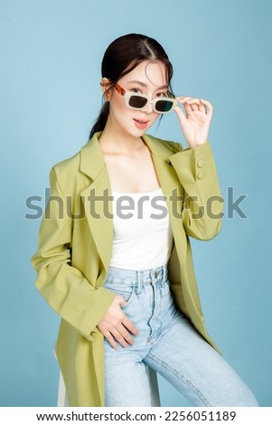 Young beautiful attractive Asian woman in casual green fashion jacket and sunglasses sitting on chair isolated on pastel blue background. Female Consumerism, lifestyle concept.