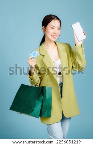 Young beautiful attractive Asian woman in casual jacket and sunglasses holding shopping bags and credit card show blank screen phone isolated on pastel blue background. Female lifestyle concept.