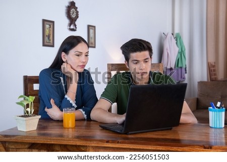 Technology Concept - Mother and son doing something in a laptop with a serious face expression. HD Image - Serious Indian mother sitting behind and looking top with a teenage boy for helping hands Royalty-Free Stock Photo #2256051503