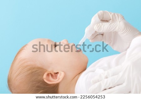 Doctor hand in rubber protective gloves holding and giving plastic tube of rotavirus vaccine on light blue background. Pastel color. Baby boy lying down and receiving medicine. Side view. Closeup. Royalty-Free Stock Photo #2256054253