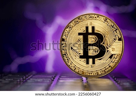 Golden metallic Bitcoin coin standing on a laptop keyboard against the background of a blurred dramatic multicolored stormy sky with lightning