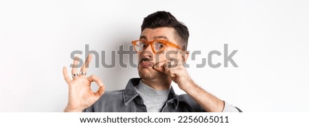 Funny young man in glasses touching his french moustache and showing okay, not bad sign, approve and like good product, standing on white background.