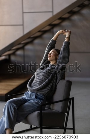 Happy millennial Caucasian woman sit in cozy chair in design home stretch relax breathe fresh air. Smiling young female renter or tenant rest in armchair relieve negative emotions enjoying weekend. Royalty-Free Stock Photo #2256070903