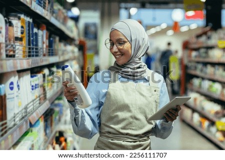 A young Arab woman in a hijab works in a supermarket, salesperson, consultant, standing with a folder in his hands in the household department, holding shampoo, making a list of products, smiling.