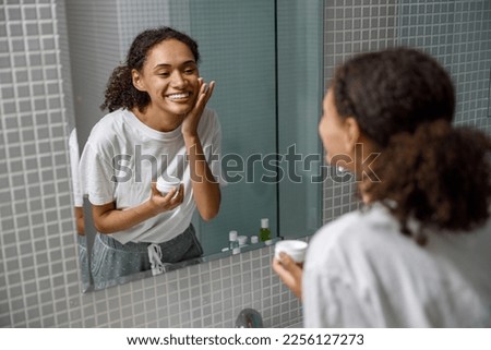 Smiling woman applying anti-wrinkle cream standing behind mirror in home bathroom. Beauty procedure Royalty-Free Stock Photo #2256127273