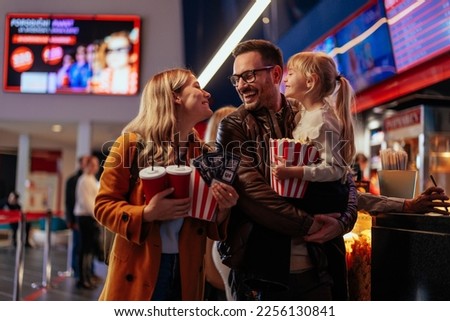 A young Caucasian family is at the movie theater, bonding as they're ready to see a movie together. The father is carrying their child and the mother is carrying popcorn, tickets and beverages. Royalty-Free Stock Photo #2256130841