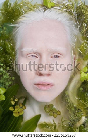 Above view of face of young serene albino woman lying in pure water with green plants while taking relaxing bath during photo session