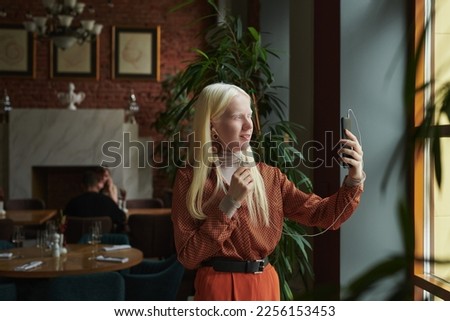 Happy young albino woman with earphones looking at mobile phone screen while standing in cafe and talking in video chat